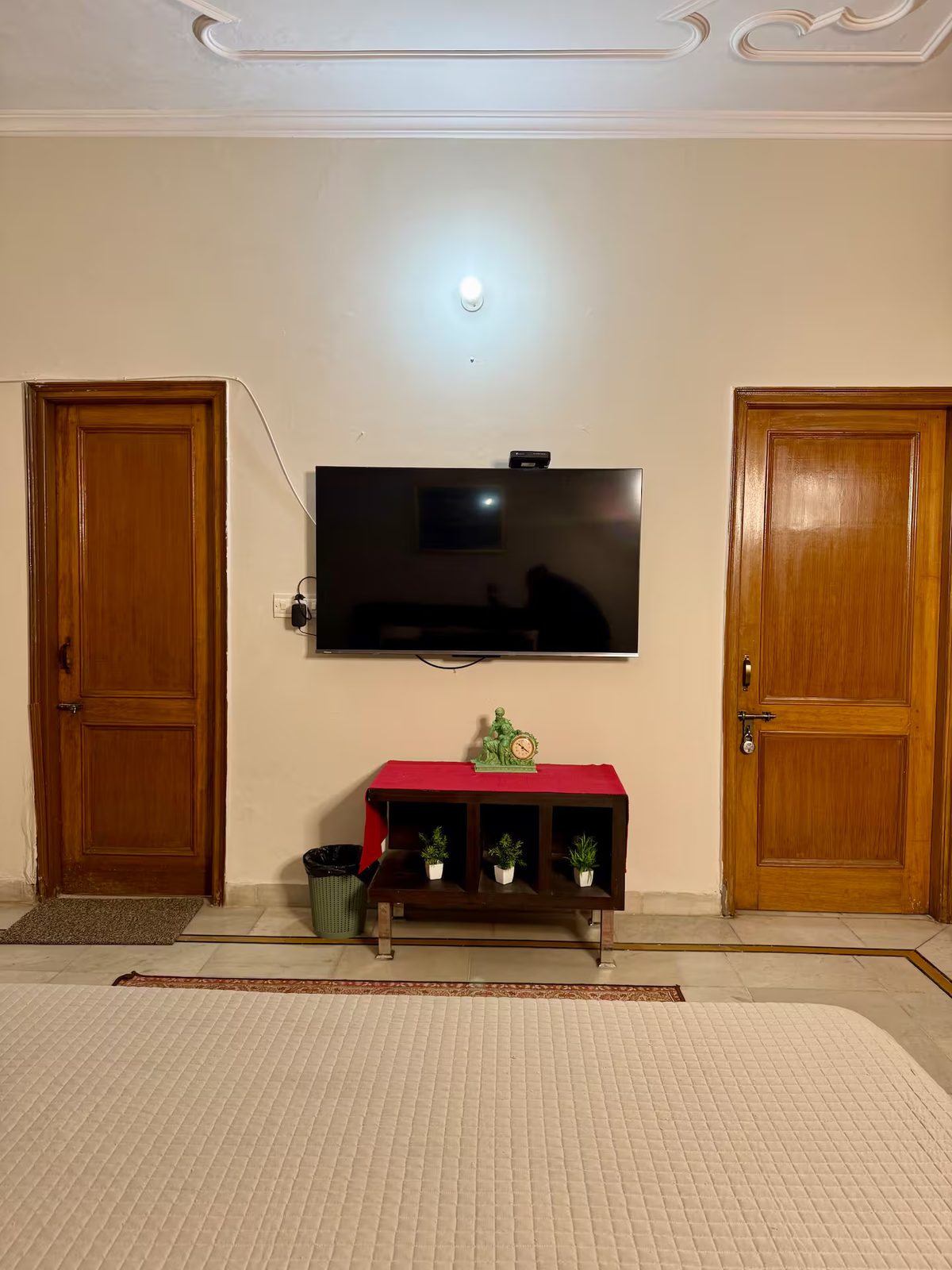 Bedroom with a large wooden bed, white quilt and pillows, bedside table, floor-to-ceiling curtains, wall-mounted air conditioner, floral artwork, wall sconce, and patterned rug.