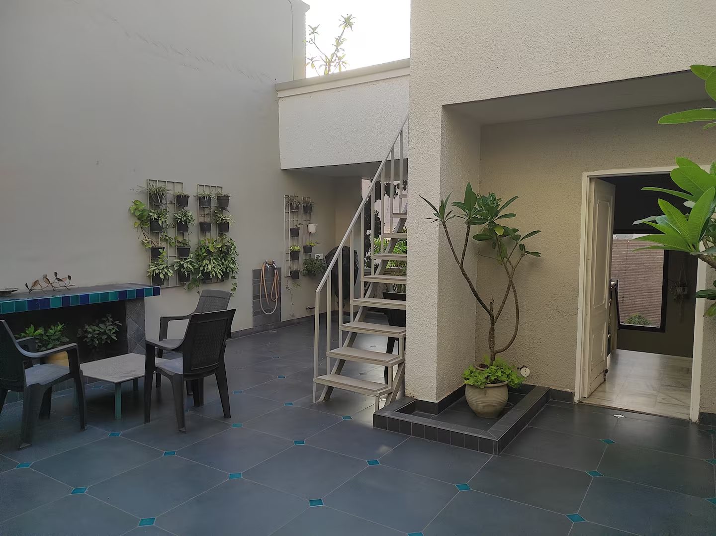 An outdoor courtyard with a table and chairs, potted plants, wall-mounted planters, tiled flooring, a white metal staircase, and an open doorway leading indoors.