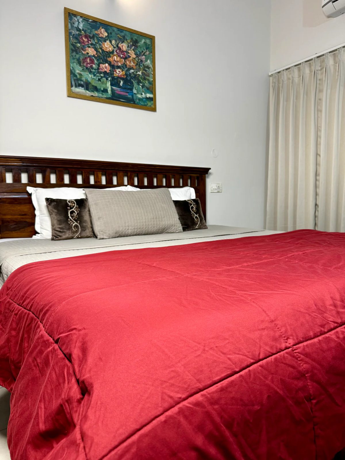 Bedroom with a wooden bed, light quilt, curtained window, built-in wooden wardrobe, wall-mounted TV, wooden console with red runner and plants, patterned rug, and tiled floor.