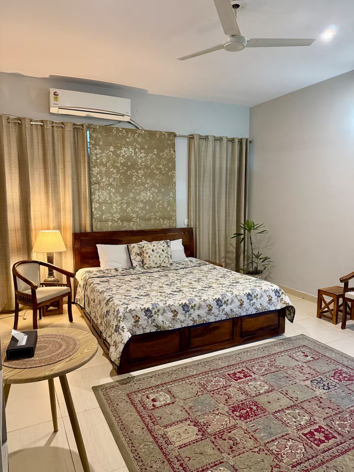 Bedroom featuring a wooden bed with white and gray bedding, patterned rug, armchair, bedside lamp, built-in shelves with books and decor, framed artwork, curtains, and a wall-mounted air conditioner.