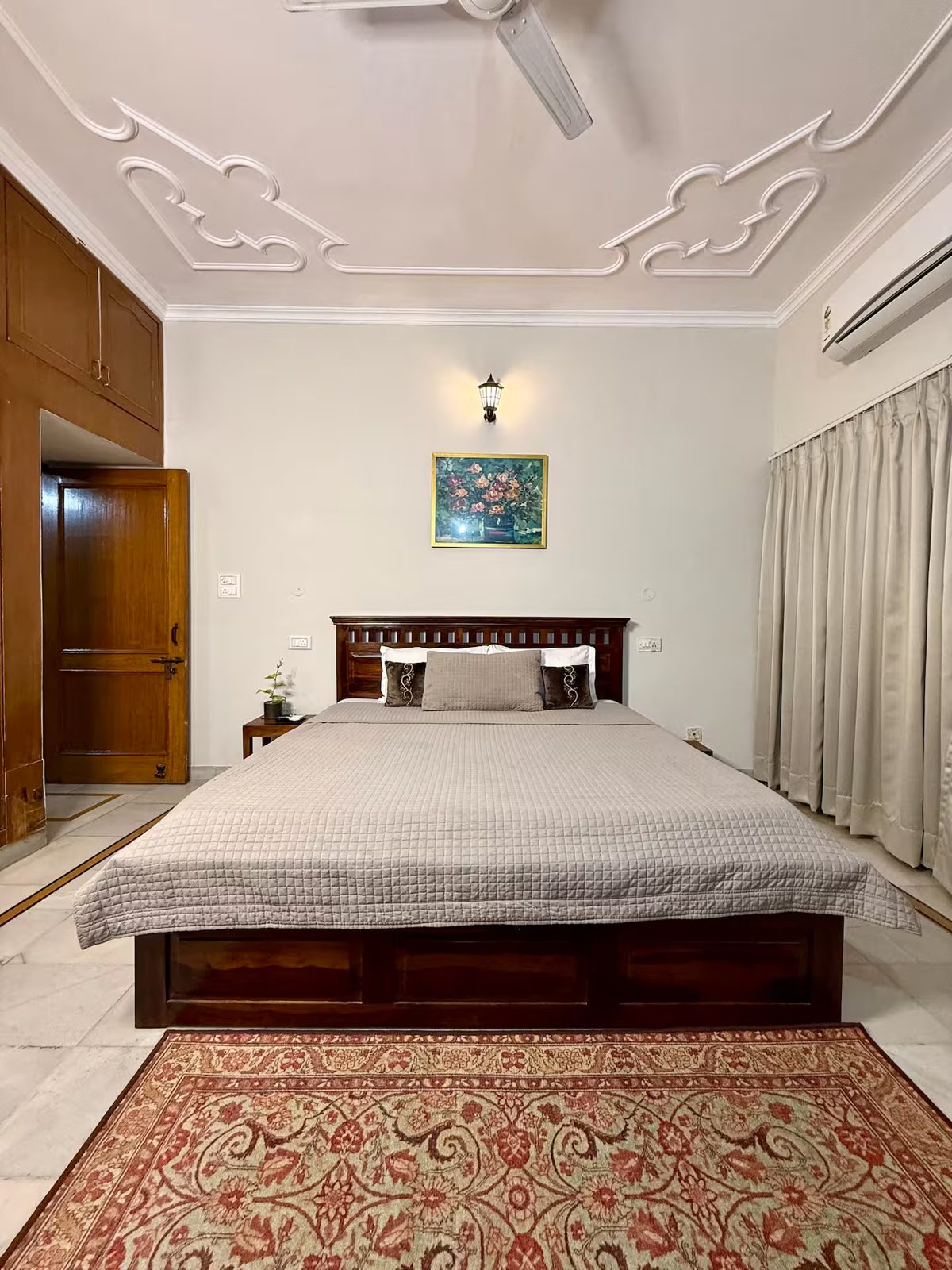 Bedroom with a wooden bed, white quilt and pillows, bedside tables, floor-to-ceiling curtains, wall-mounted air conditioner, framed artwork, and decorative ceiling molding.