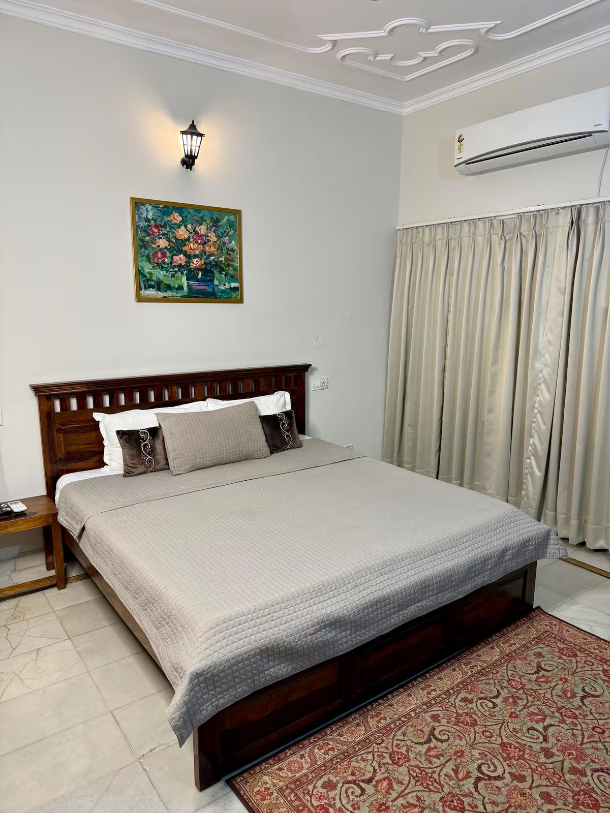 Bedroom with a large wooden bed, white quilt and layered pillows, built-in wooden wardrobes, a bedside table with a potted plant, framed floral artwork, and decorative ceiling molding.