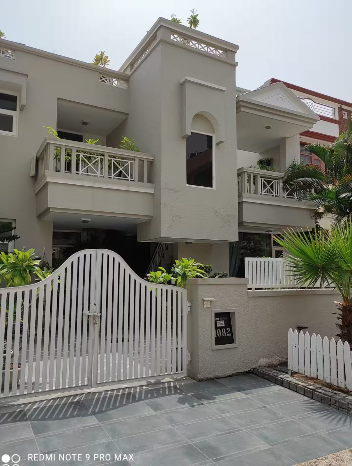 A multi-story villa with white walls, balconies, a covered entrance, white metal gate, tiled driveway, and landscaped plants along the front boundary.