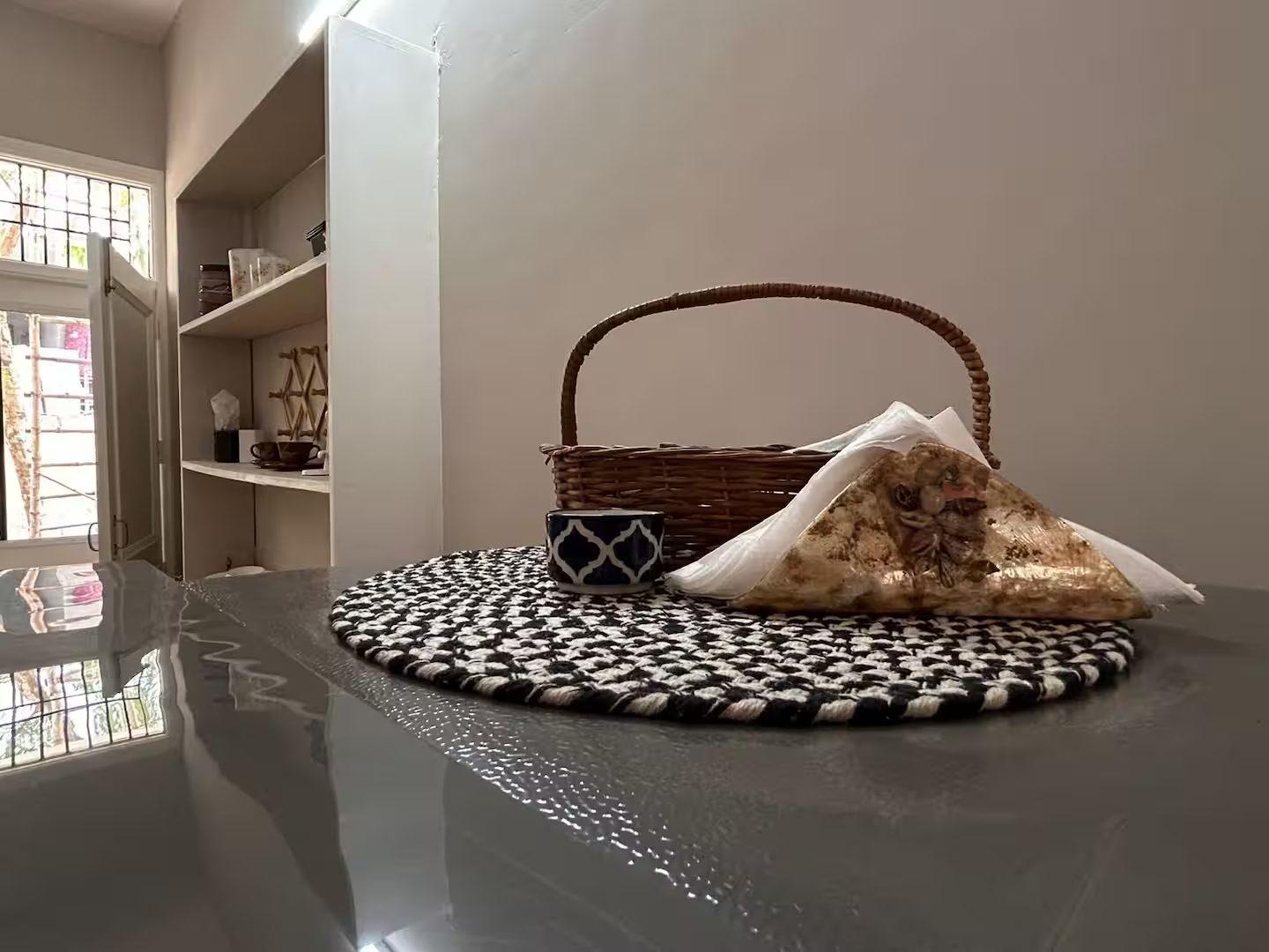 Dining table with a black-and-white woven placemat, a patterned cup, a wicker basket, and folded napkins. Built-in shelving, a doorway, and windows are visible in the background.