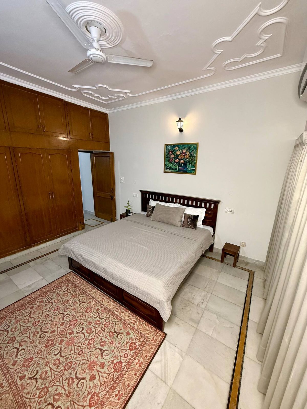 Bedroom with a wooden bed, red quilt, white and brown pillows, wooden wall cabinets, floral painting, wall light, tiled floor, and an open wooden door.