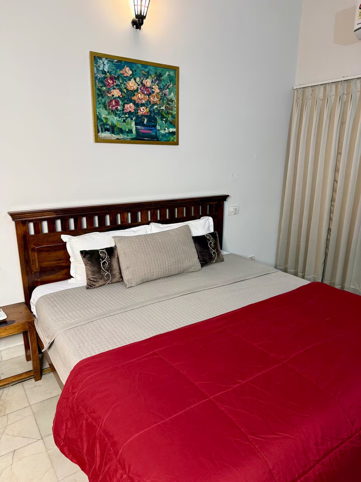 Bedroom showing a wooden bed with a red quilt, white and brown pillows, floral wall painting, floor-length curtains, and a wall-mounted air conditioner.