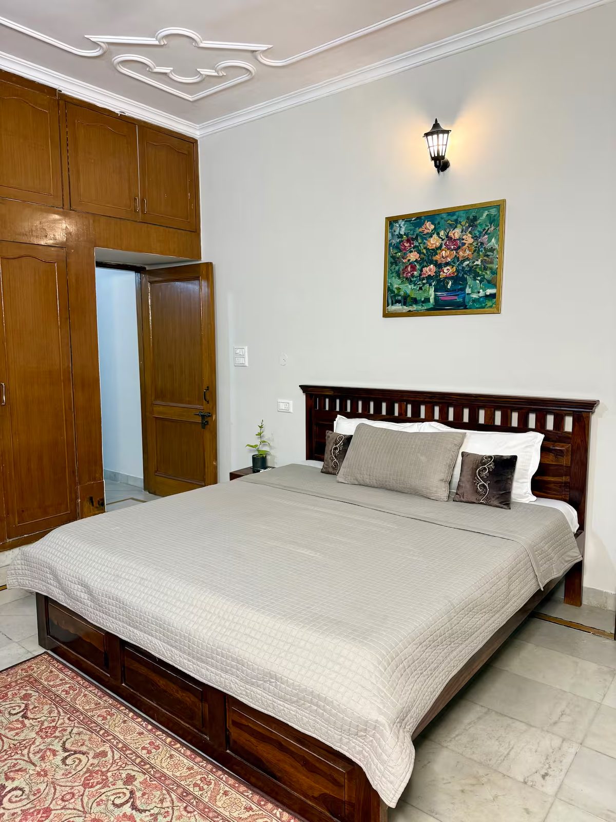 Close-up of a wooden bed with a white quilt and layered pillows, beside a wooden nightstand holding a potted plant and remote controls. Full-length curtains cover the window behind the bed.