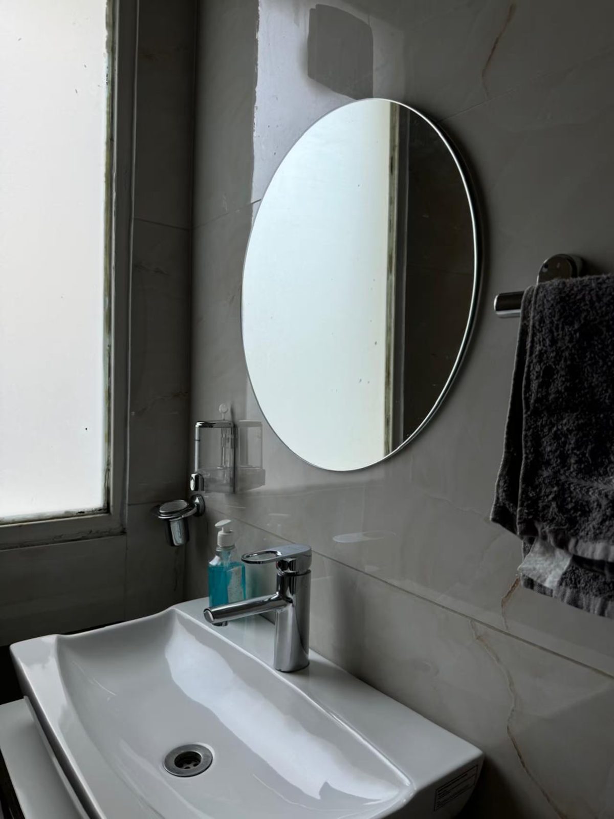 Bathroom vanity with a white rectangular sink, chrome faucet, round wall mirror, soap dispenser, towel holder, and a window providing natural light.