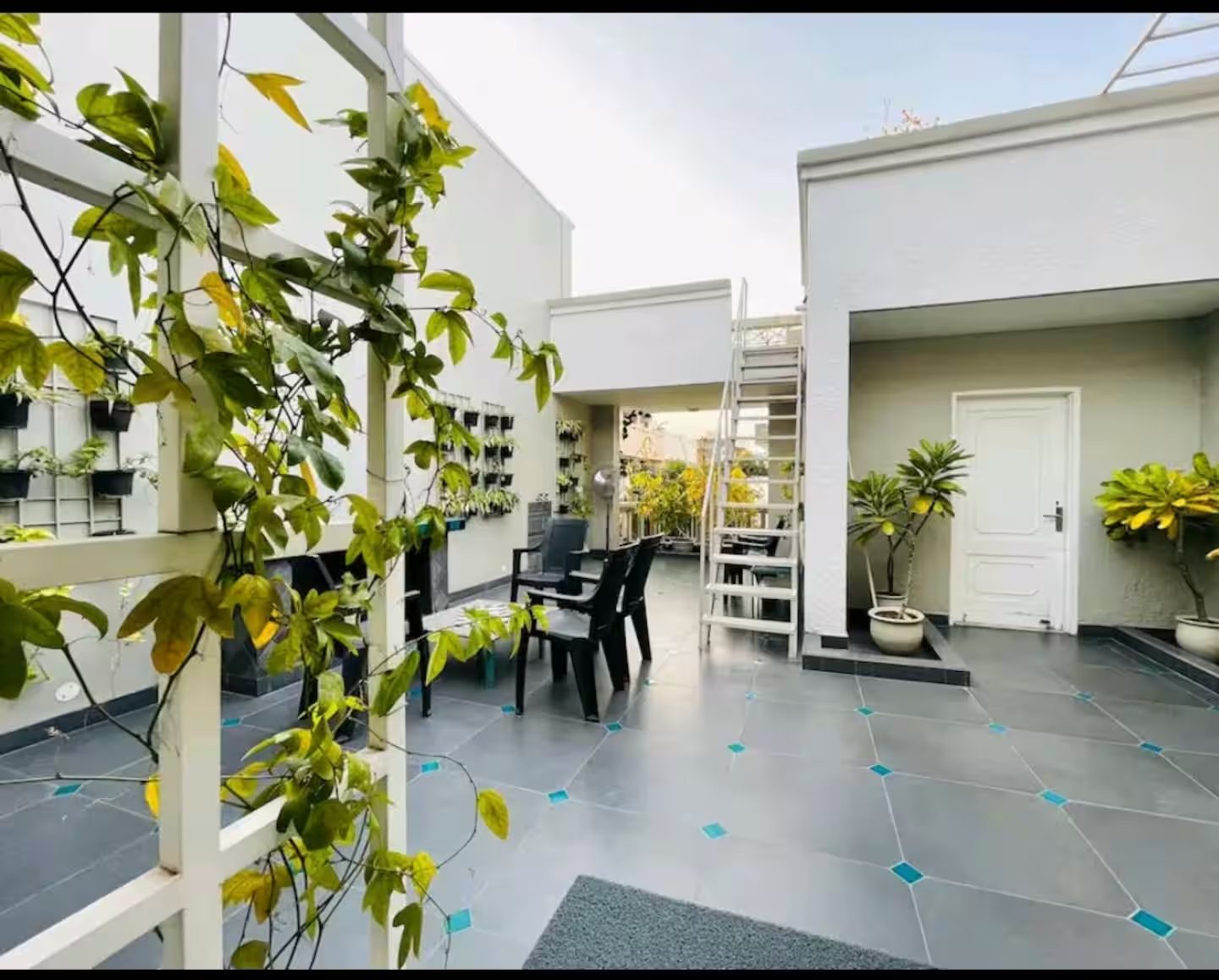 An outdoor terrace with tiled flooring, potted plants, black chairs, a vertical garden, a white staircase, and a covered doorway.