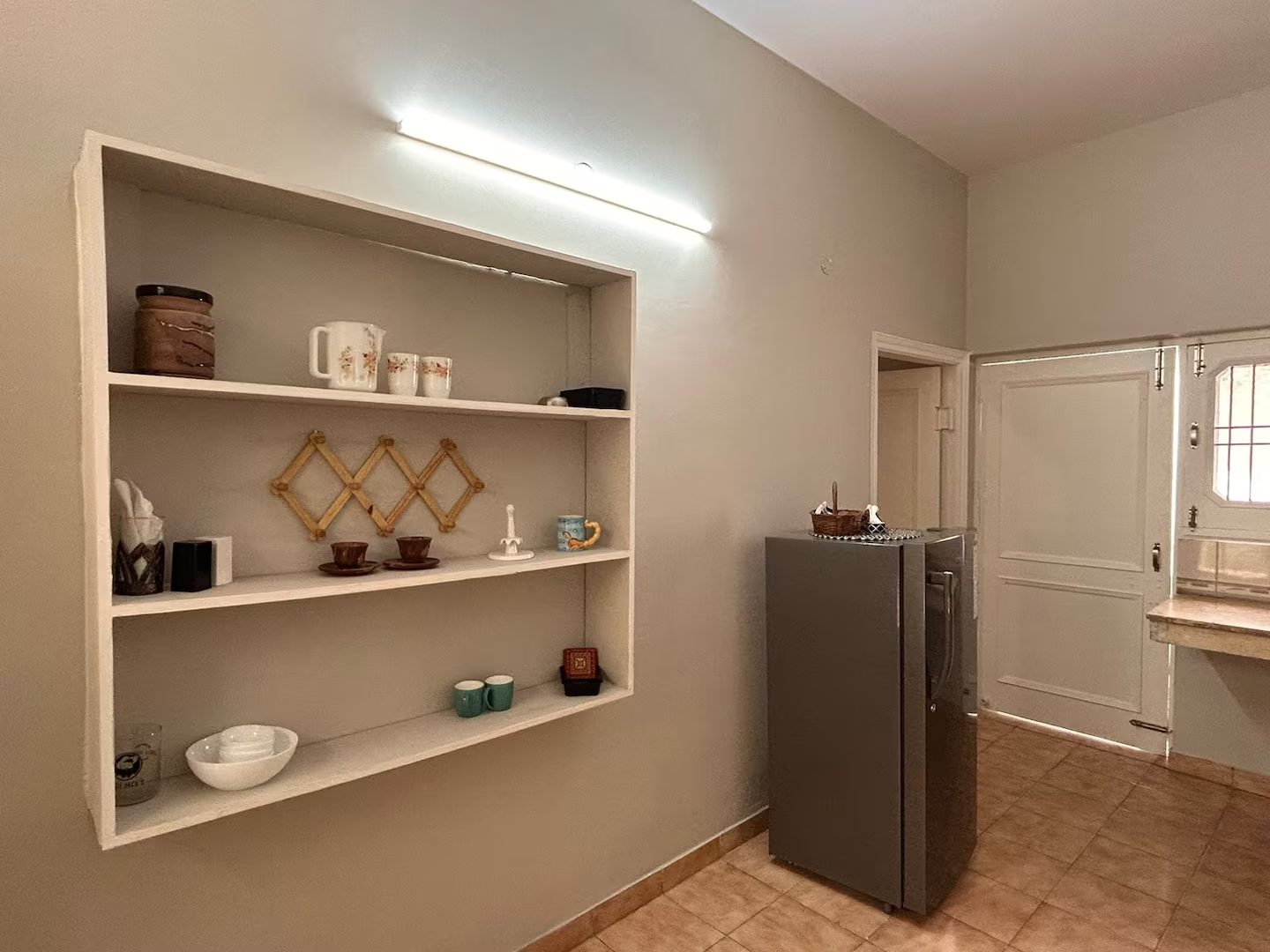 Kitchen area with open shelving displaying cups, bowls, dishes, and decorative items, a freestanding refrigerator, tiled floor, white doors, and a countertop with a window visible on the right.