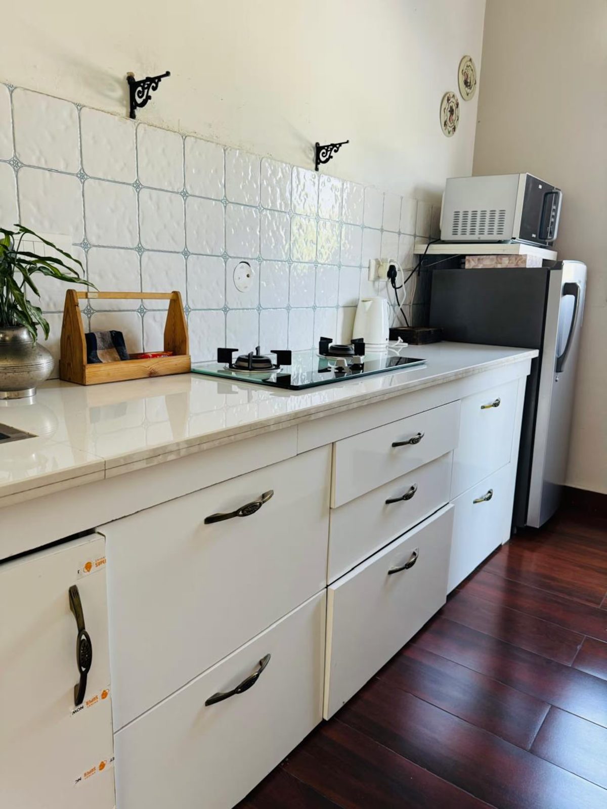 Kitchen with white cabinets and tiled backsplash, two-burner gas cooktop, microwave, electric kettle, refrigerator, countertop storage, and dark wood flooring