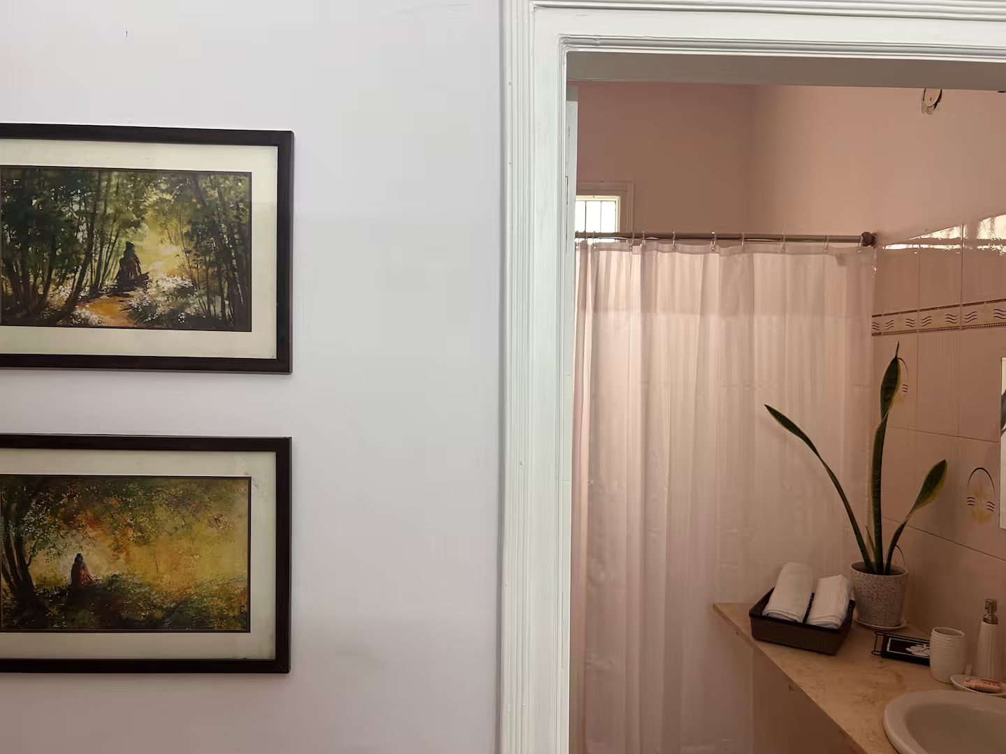 Bathroom with a white sink and chrome faucet, large wall mirror, patterned tiled walls, white shower curtain, potted plant, folded towels, toiletries, and a window with metal bars.