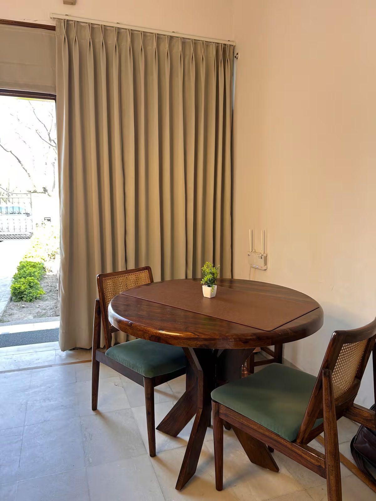 Dining area with a round wooden table, two wooden chairs with green cushions, floor-to-ceiling curtains, and a glass door opening toward the garden.