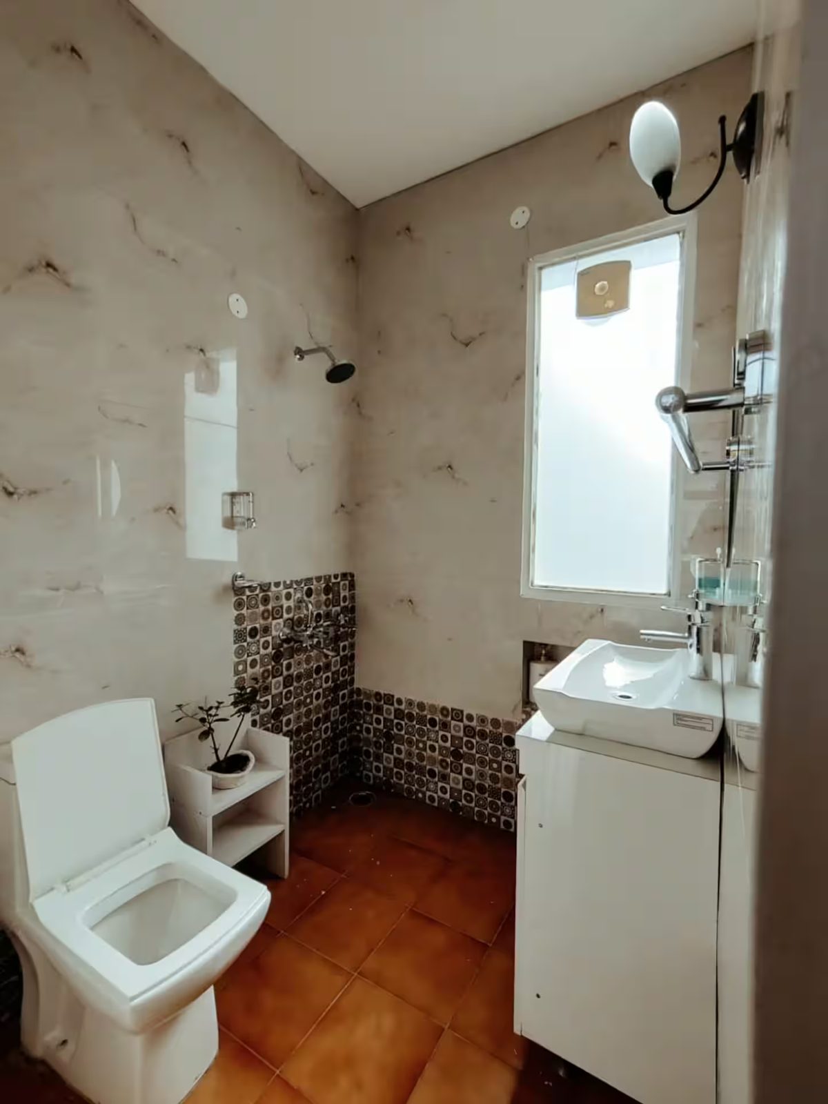 A bathroom with a toilet, vessel sink on a vanity, glass-enclosed shower, wall-mounted fixtures, frosted window, patterned wall tiles, and brown floor tiles.