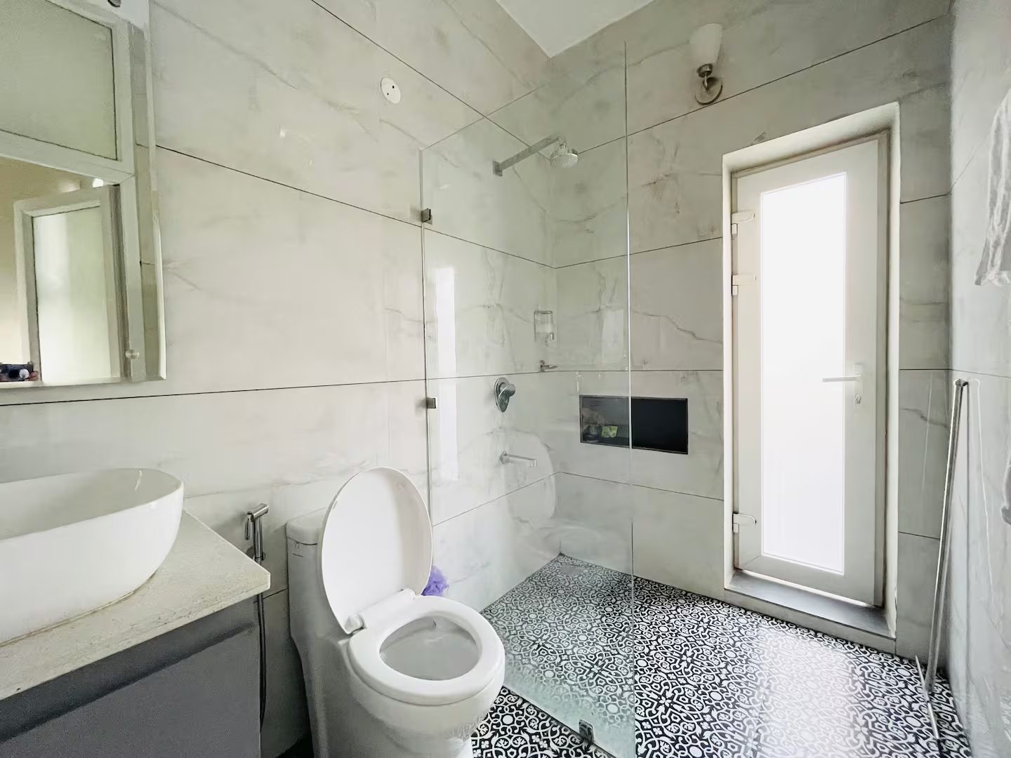 A bathroom with white marble-patterned wall tiles, a glass-enclosed shower, wall-mounted showerhead, white toilet, vessel sink, patterned floor tiles, mirror, and a frosted glass door.