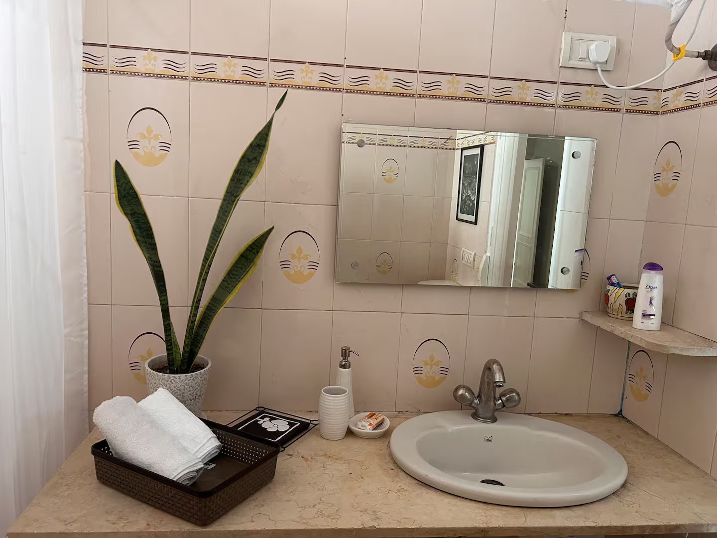 Bathroom vanity with a round white sink, chrome faucet, rectangular wall mirror, patterned ceramic tile walls, soap dispenser, toiletries, folded towels, and a potted snake plant.