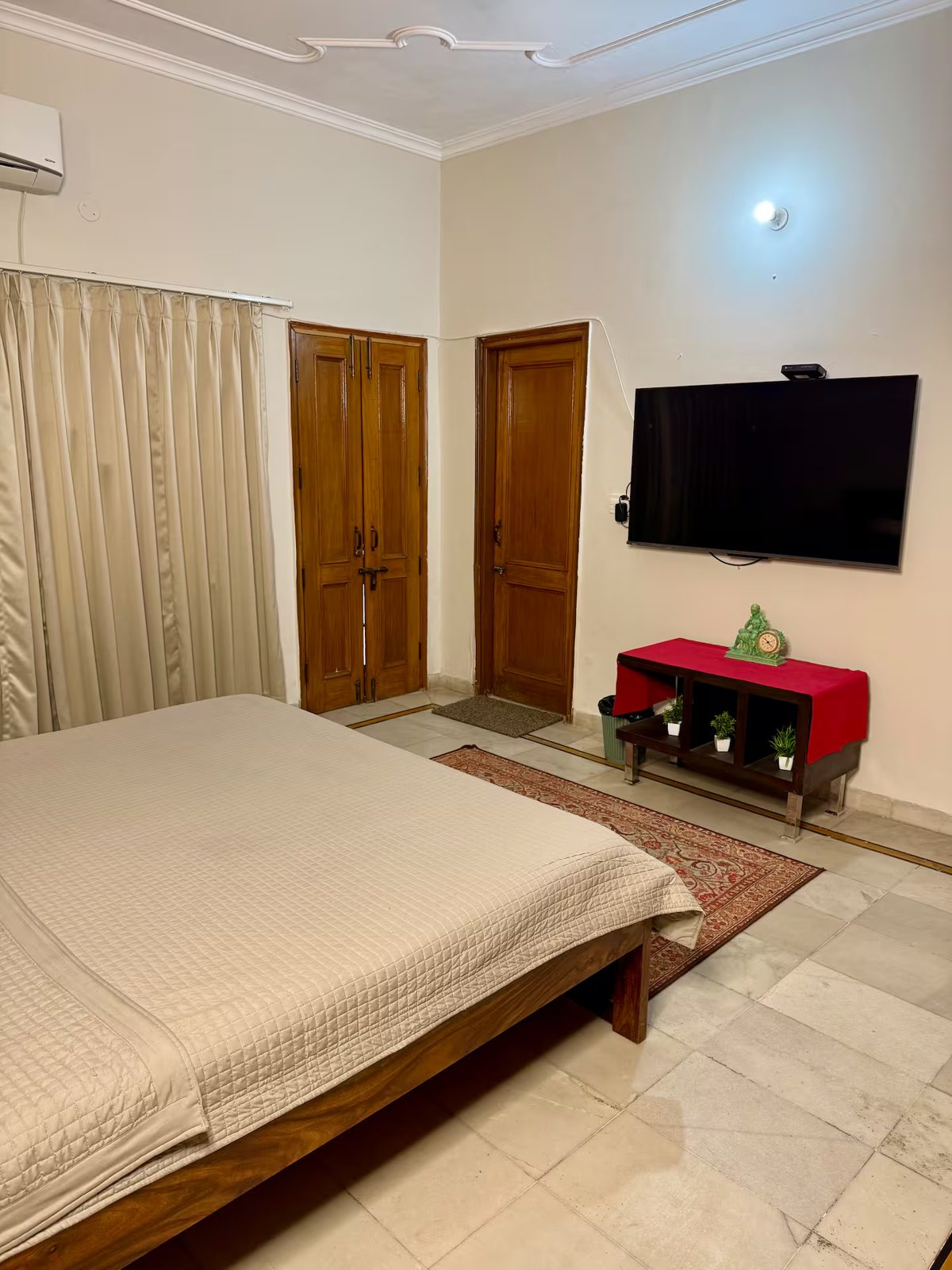 Bedroom with a wooden bed, light quilt, curtained window, built-in wooden wardrobe, wall-mounted TV, wooden console with red runner and plants, patterned rug, and tiled floor.