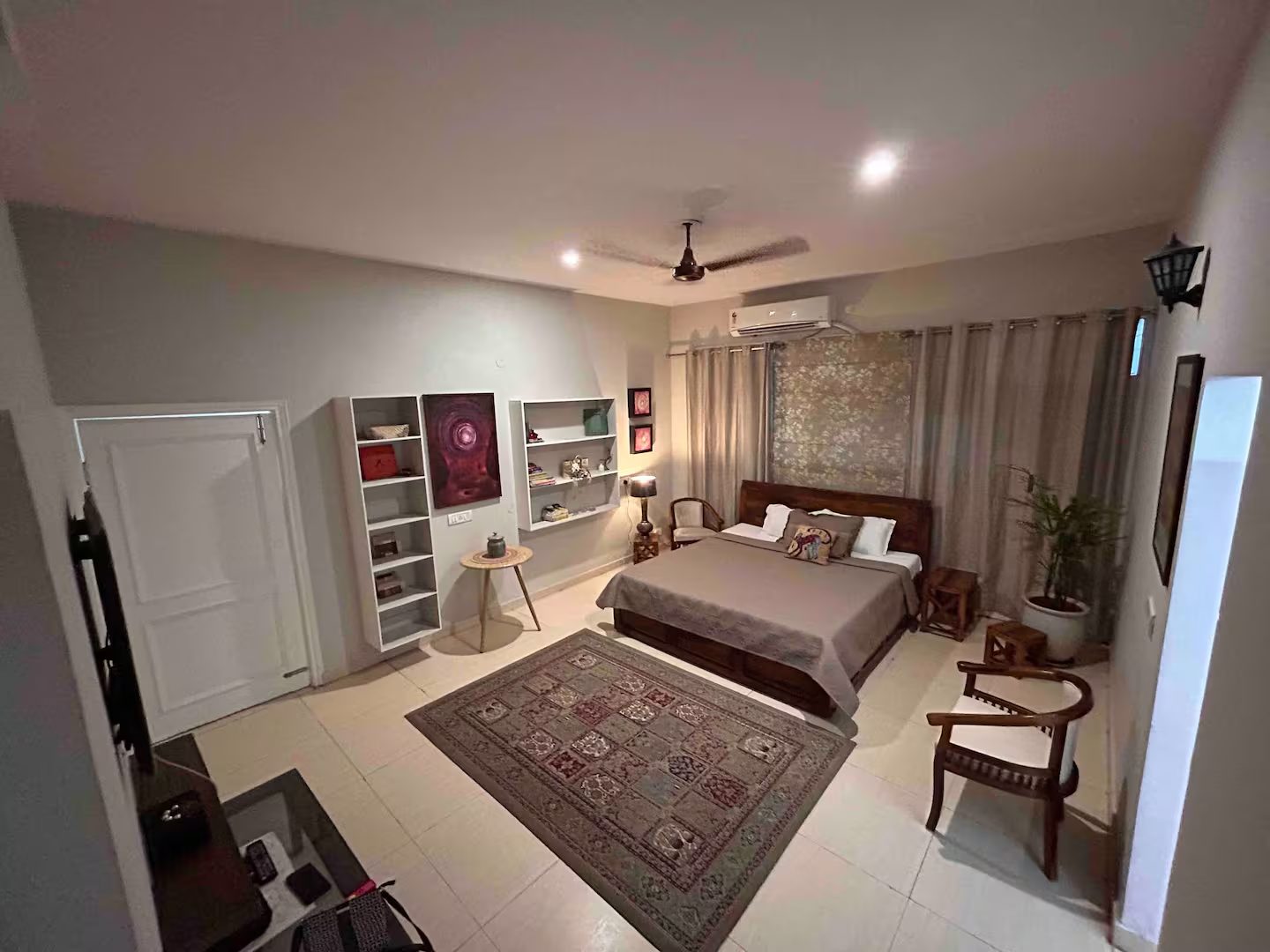 Bedroom featuring a wooden bed with patterned bedding, decorative pillows, bedside table, floral wall panel, curtains, ceiling fan, wall-mounted air conditioner, and a windowed door.