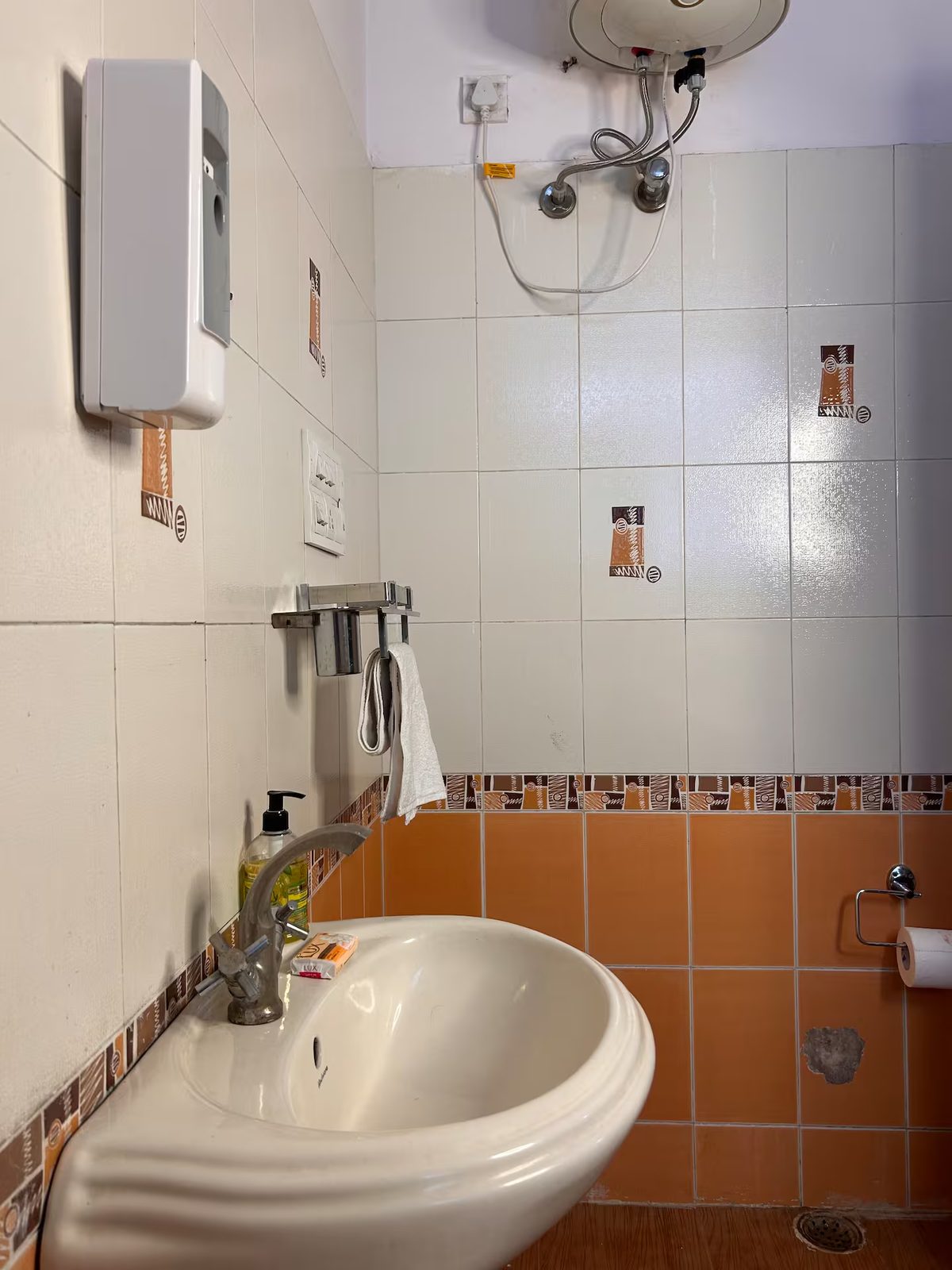 Bathroom with tiled walls, a wall-mounted water heater, toilet, partial sink, towel rack, toilet paper holder, floor drain, and orange-and-white patterned tilework.