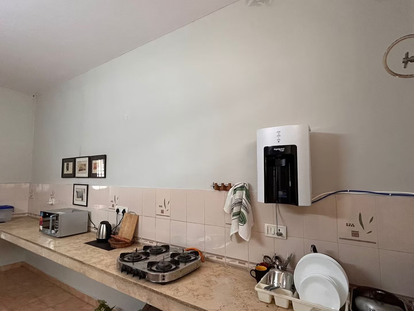 Kitchen counter with a four-burner gas stove, microwave, electric kettle, wall-mounted water dispenser, toaster, dish rack with plates, utensils, cutting board, and tea towel.