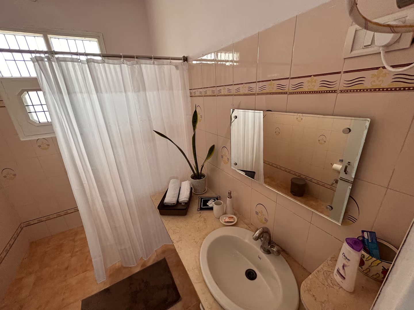 Bathroom with a white sink and chrome faucet, large wall mirror, patterned tiled walls, white shower curtain, potted plant, folded towels, toiletries, and a window with metal bars.