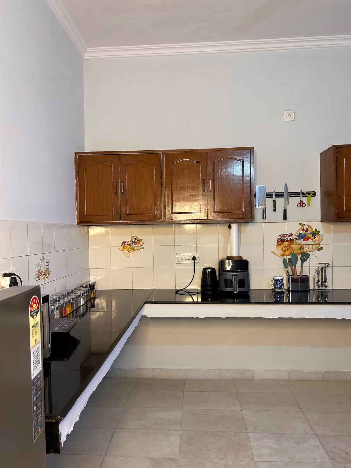 Kitchen with L-shaped black countertops, wooden wall cabinets, a microwave, air fryer, electric kettle, spice jars, utensil holders, and wall-mounted kitchen tools.