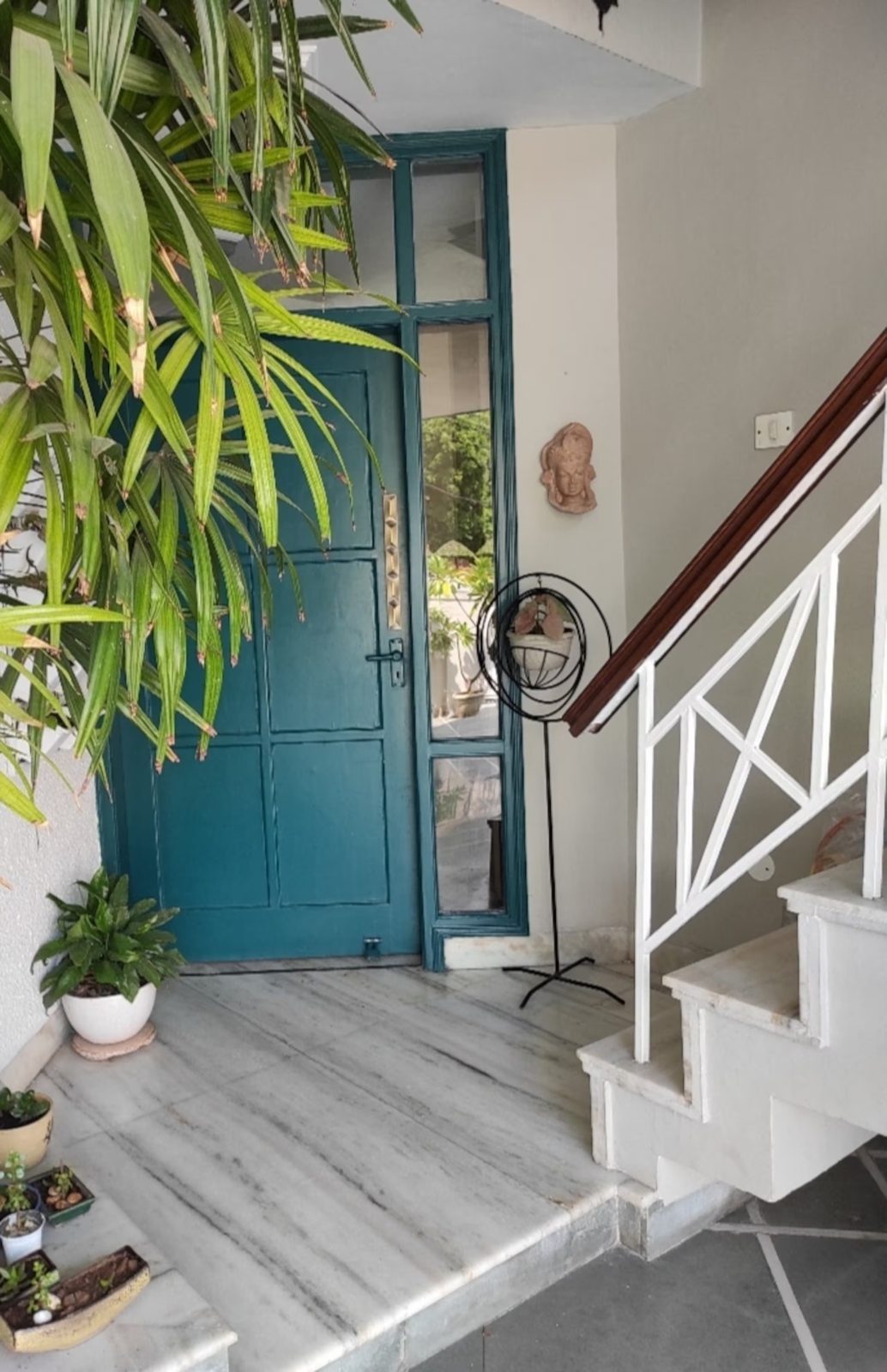 Entryway with a teal door and adjacent glass panels, marble flooring, white staircase railing, potted plants, and a decorative wall sculpture.