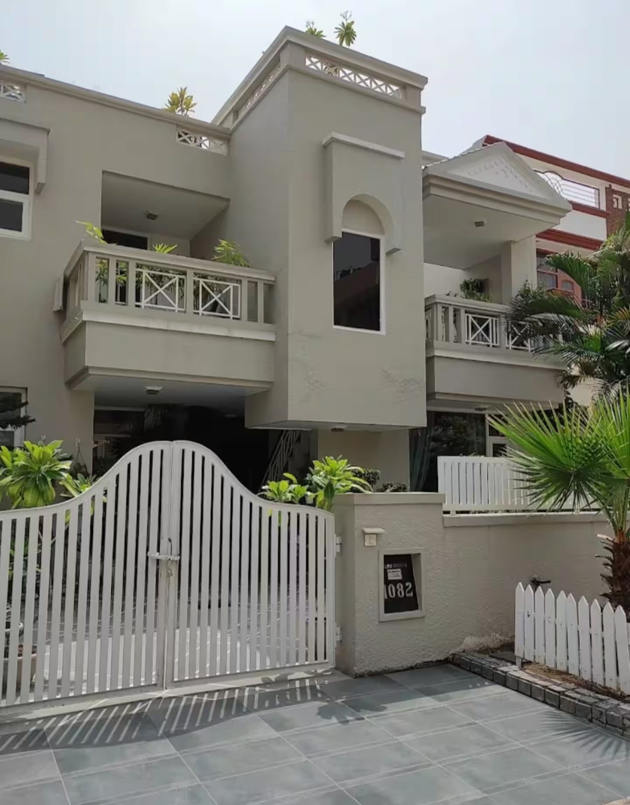 The villa's front exterior with a white metal gate, balconies with white railings, large windows, tiled driveway, and landscaped plants along the entrance.