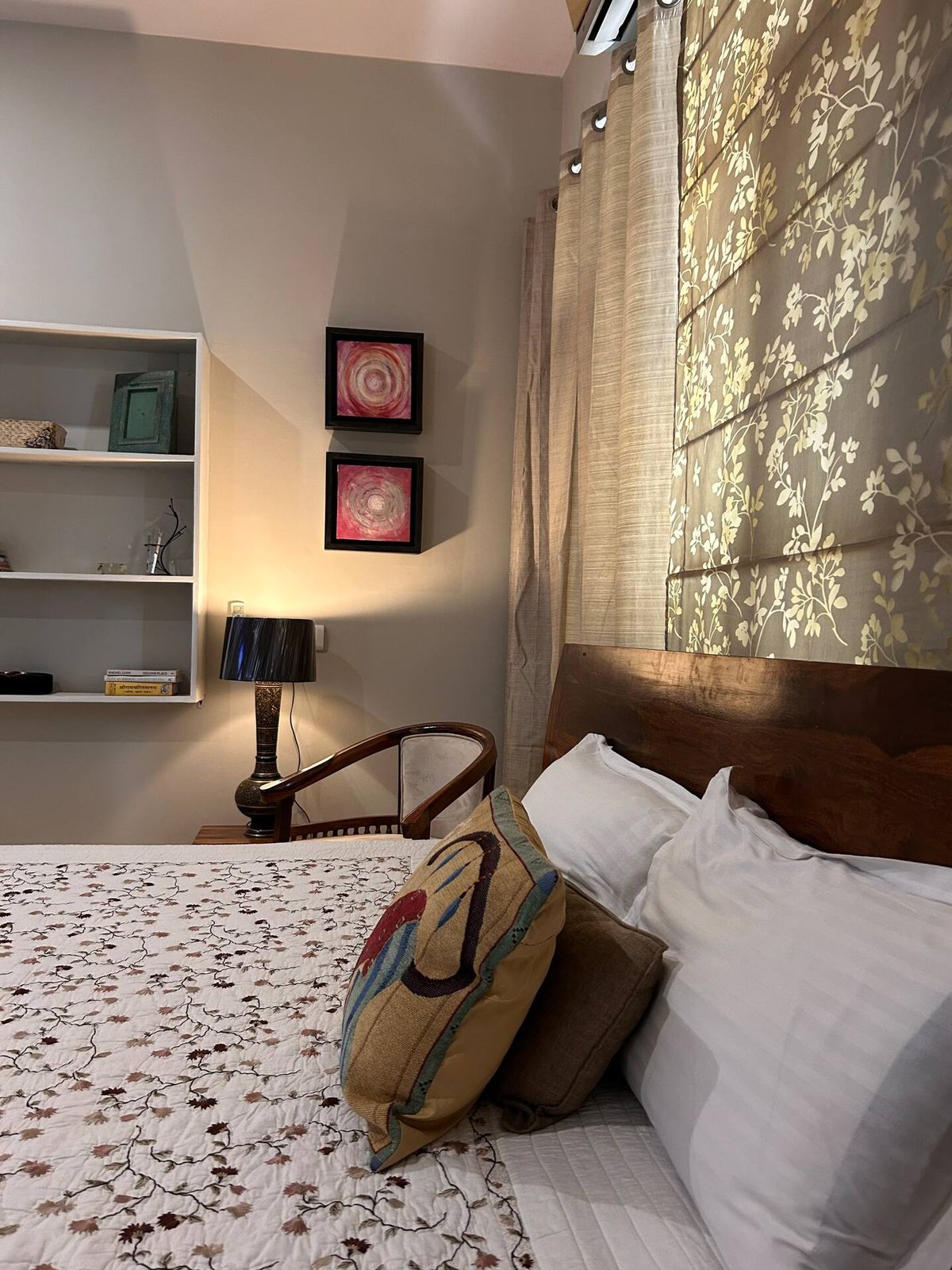 Bedroom with a wooden bed, patterned bedspread, decorative pillows, armchairs, bedside tables, large patterned rug, wall shelving, framed artwork, curtains, ceiling fan, air conditioner, and a potted plant.